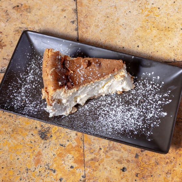 A slice of cheesecake with a brown crust and creamy white filling, dusted with powdered sugar.