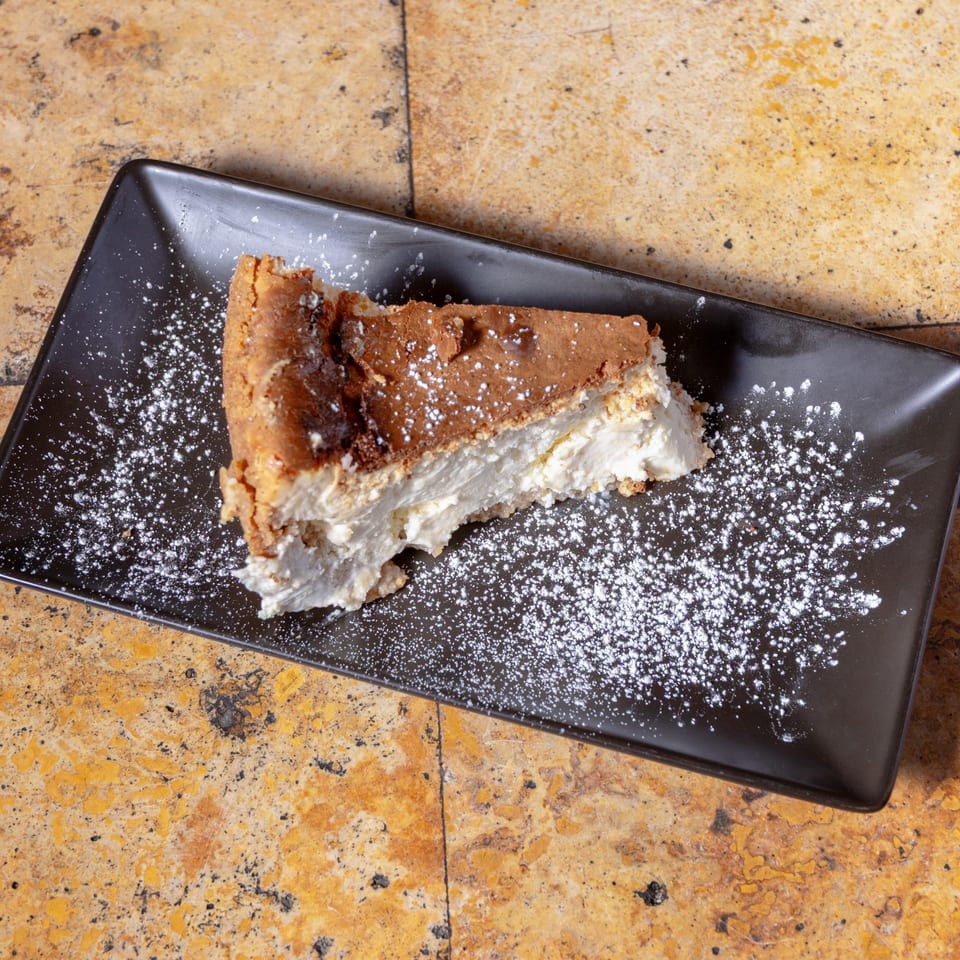 A slice of cheesecake with a brown crust and creamy white filling, dusted with powdered sugar.