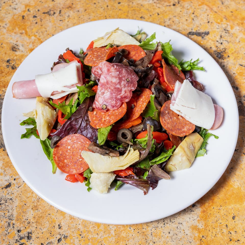 A white plate holds a vibrant Italian antipasto salad with meats, cheeses, artichokes, and olives.