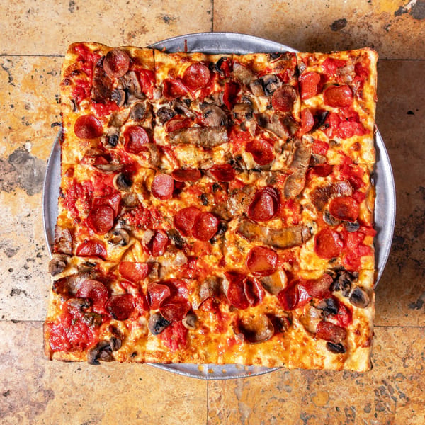 Square Carmelas Brick Oven Pizza with pepperoni, sausage, and mushrooms on a metal tray.