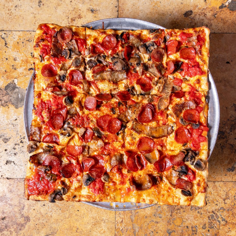 Square Carmelas Brick Oven Pizza with pepperoni, sausage, and mushrooms on a metal tray.