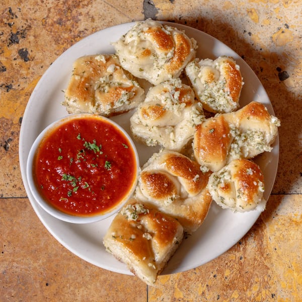 A white plate of golden-brown garlic knots with a side of red marinara sauce, garnished with parsley.