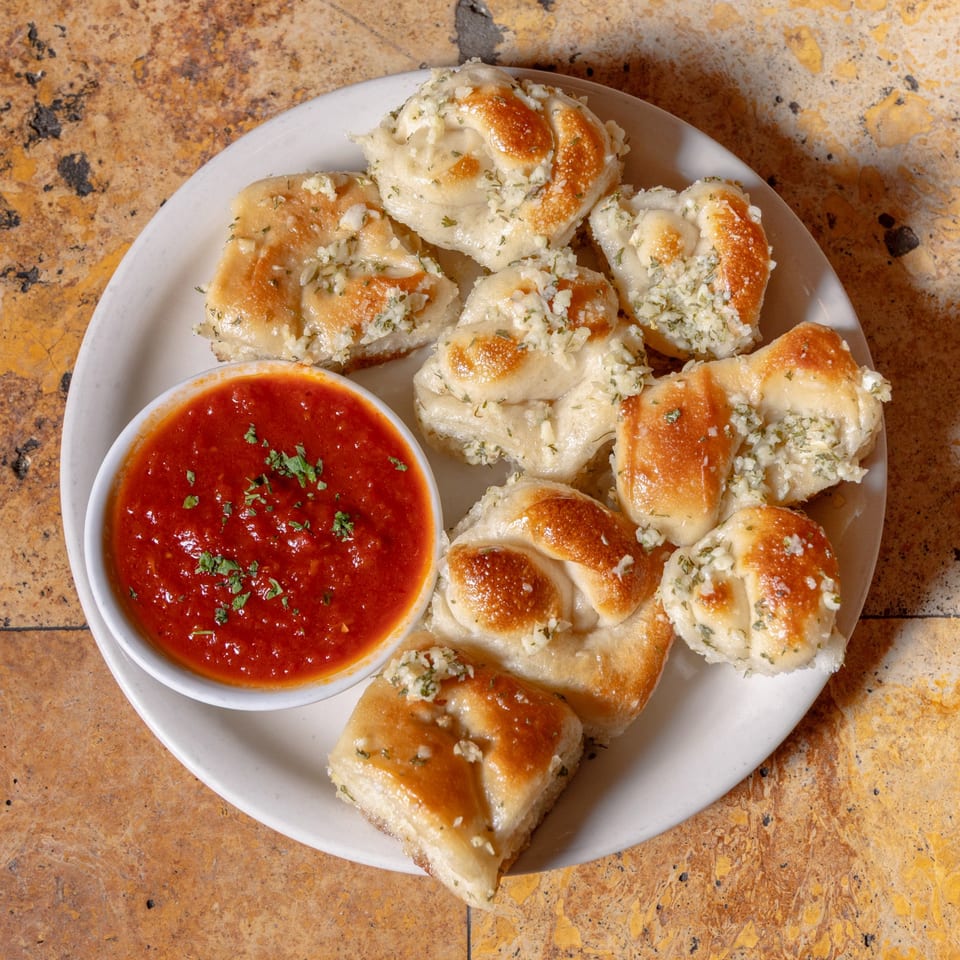 A white plate of golden-brown garlic knots with a side of red marinara sauce, garnished with parsley.