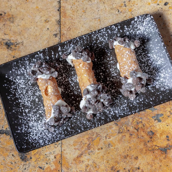 Three cannolis with white cream filling and chocolate chips, dusted with powdered sugar on a black plate.